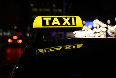 Ein beleuchtetes gelbes Taxischild auf einem Fahrzeugdach, im Hintergrund Lichter einer Stadt, aufgenommen bei Nacht